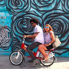 Two people riding a bicycle together against a vibrant blue mural